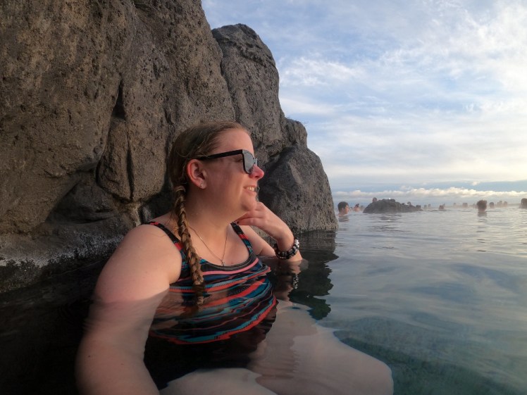 A selfie at the Sky Lagoon. I'm sitting against the cliff wall, facing the sunset, rune pendant and tiny huggie hoop earrings visible.