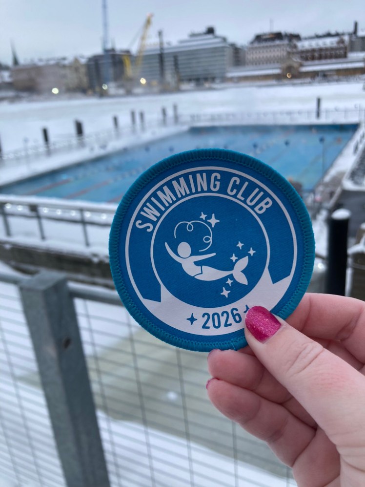 A blue Swimming Club 2026 patch with a mermaid in the middle of it, held up in front of the pool.