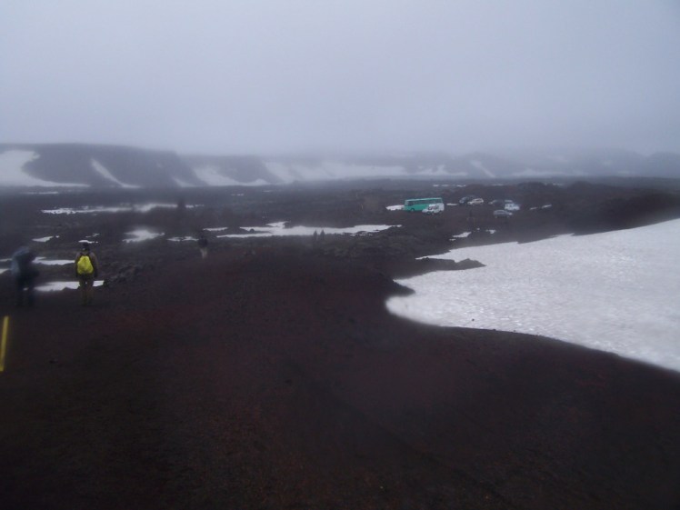 Walking across Askja. It's very cloudy, hanging low across the mountain, but my camera lens being wet & muddy is not helping. The mountain is a deep red that's almost black, patched with snow and you can see an assortment of cars and buses parked just a little distance away.