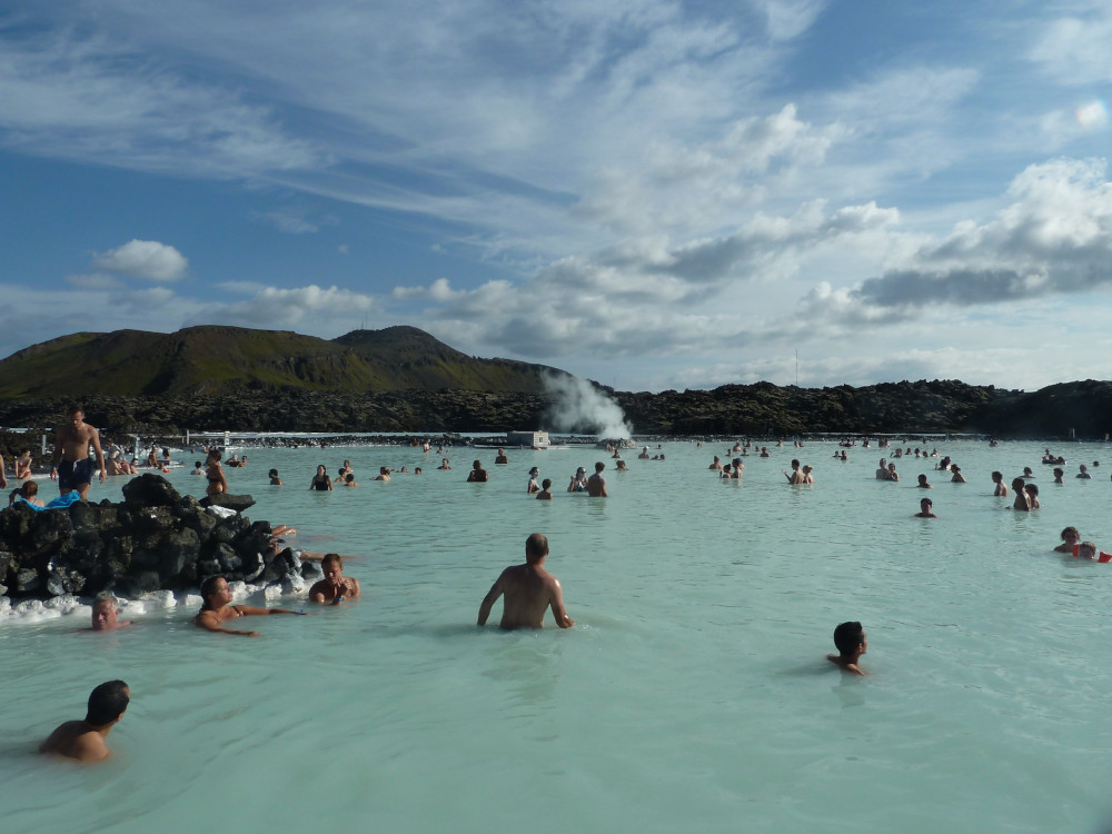 The Blue Lagoon in the summer, looking quite green in the sunlight. There are lots of people in the water and on the horizon is the power station, steaming away.