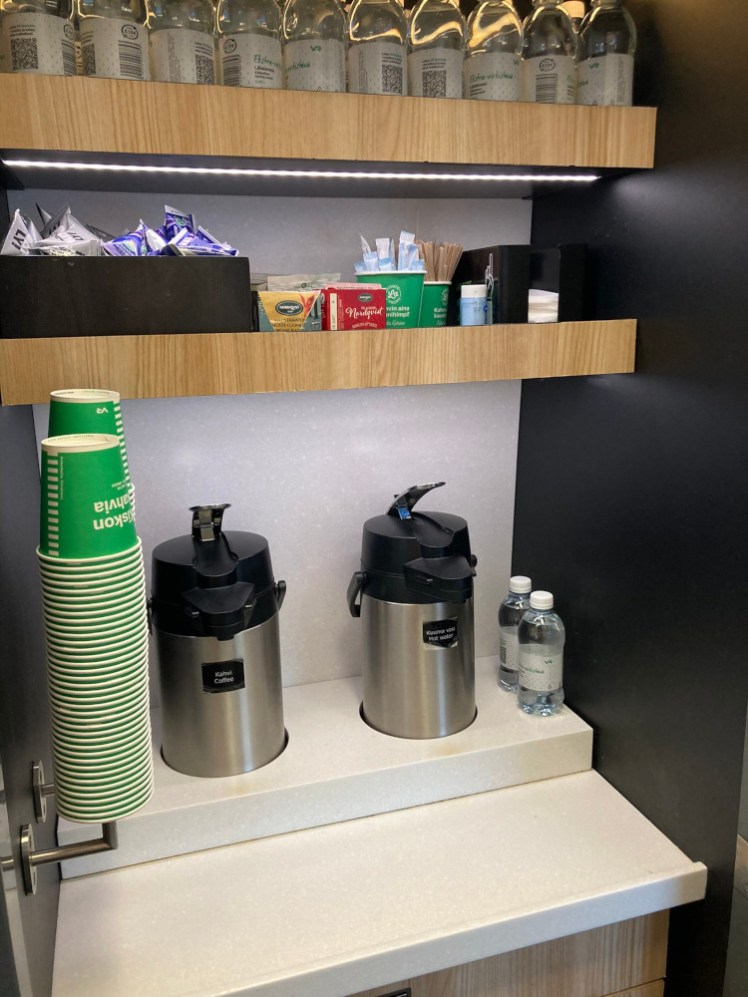 The drinks station in the middle of the Ekstra Calm carriage. There are two jugs of coffee and hot water, two stacks of green paper cups and a shelf of tea, sugar, stirrers and milk.