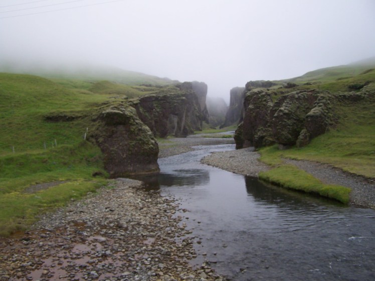 Fjaðrárgljúfur, a green canyon with a gentle river rolling through it and with a mist sitting just above it.