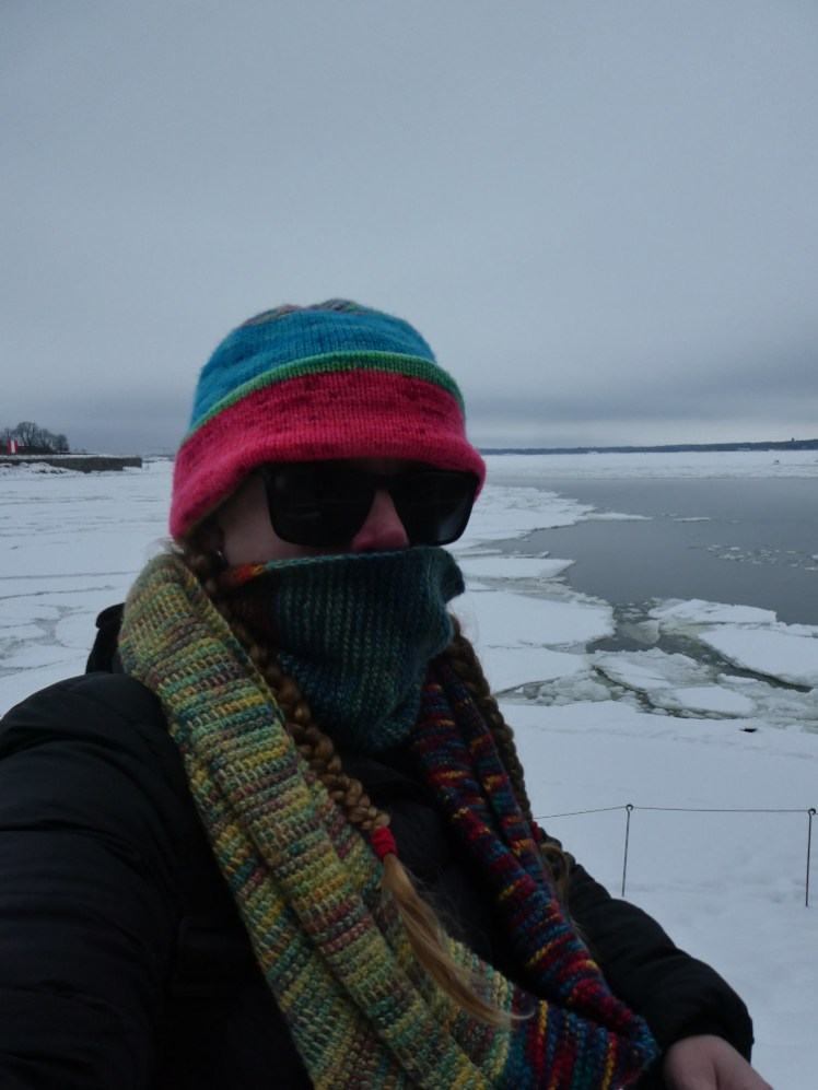 A selfie with a colourful hat and infinity scarf. One loop of it is pulled up over my face because it's so cold and because everything's so white, I'm wearing sunglasses so you can't even see me. Behind, you can see snow-covered sea ice to the left and wetter, more open sea to the right. This is from the top of the walls on Suomenlinna, not from the ferry. I didn't think of taking a selfie on the ferry, it was too cold.