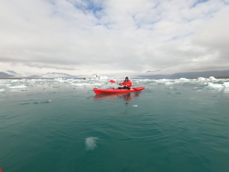 Me in a red kayak and a red suit on the background of a teal glacial lake with icebergs floating in the background.
