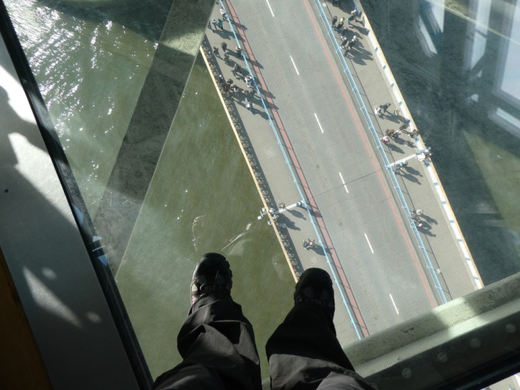 My feet kind of silhouetted on the glass floor against the road and river below, which are well lit because it's a sunny day.