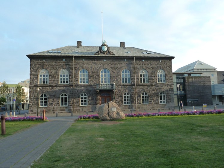 A flat-fronted two-storey building of dark stone set in front of a quiet road and a grassy bit of square.