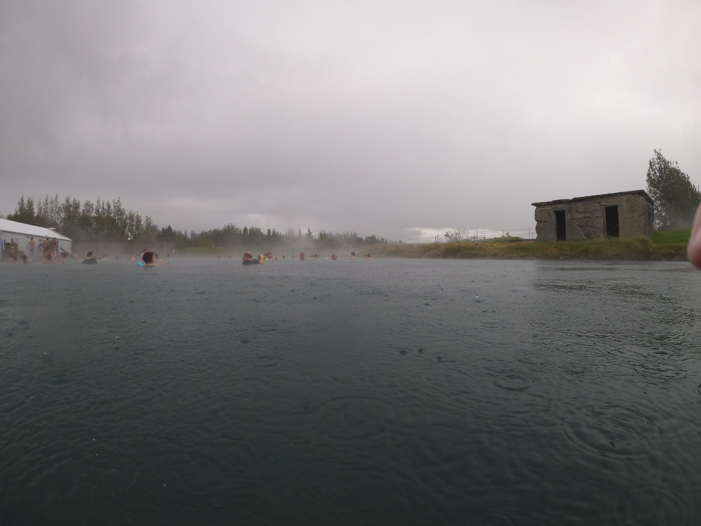 The Secret Lagoon in the rain. On the right is a roofless old hut and the field of geysers that fills the pool.