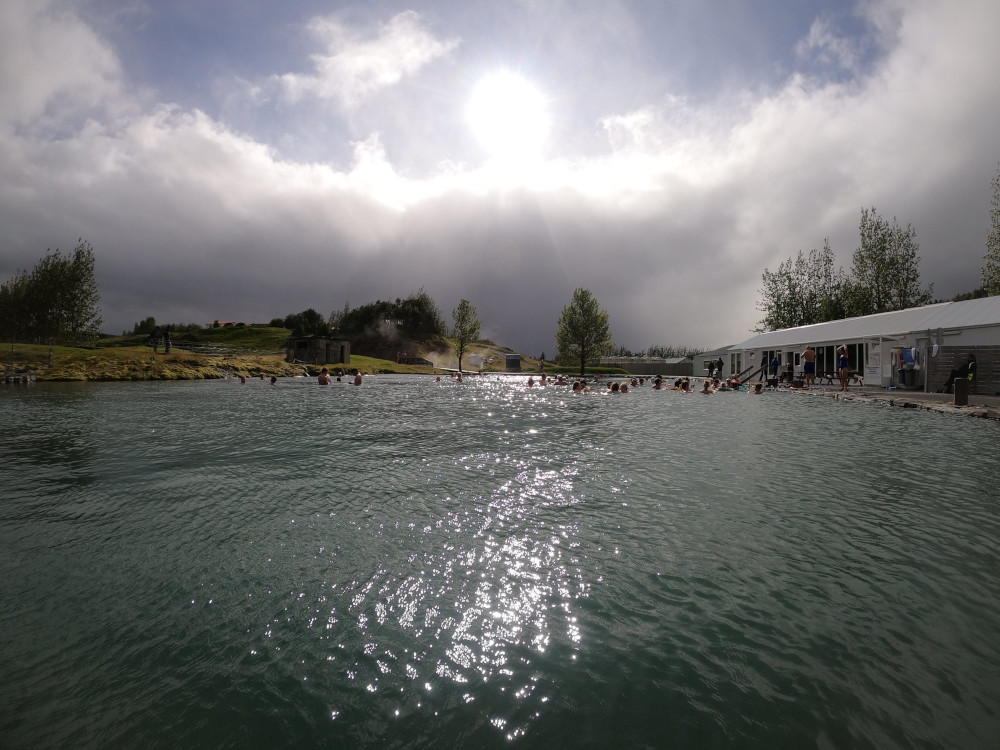 The Secret Lagoon gleaming in the sunshine, with the building on the left and the geysers on the right.