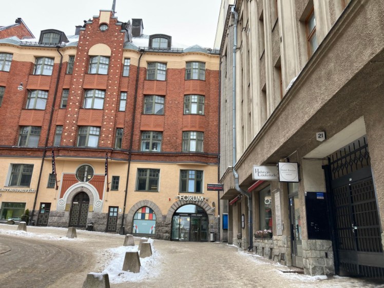 Yrjönkatu's north end, where a building in red brick and yellow plaster meets a building in a greyer Art Nouveau style. The ground is thick with yellow sand covering ice and snow.