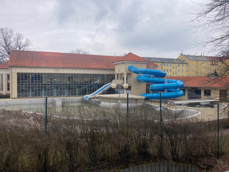 Aquaforum's outside area seen from the station. There's a slide on the left-hand side of a kind of tower attached to the back of a building and a bigger spiral slide attached to the other side and they both run down into what must be massive sparkly pools of blue water in the summer but in early April are just empty concrete vats.