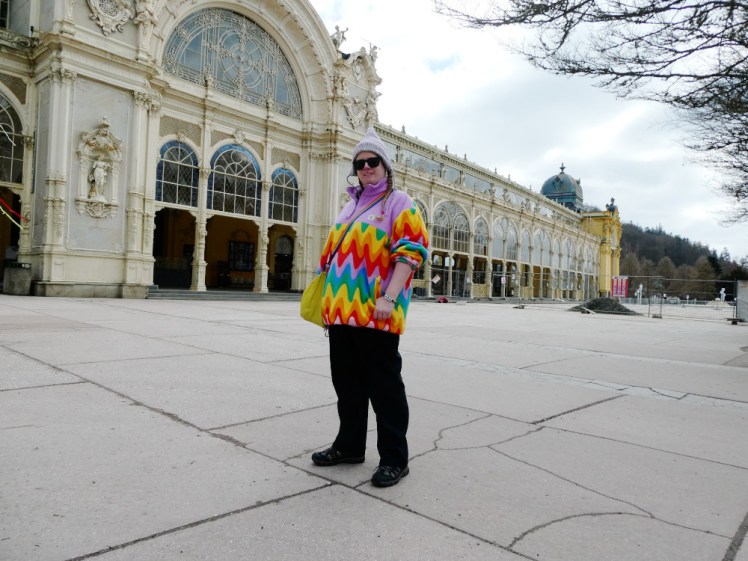 A selfie outside the Colonnades, a Victorian wrought iron covered promenade. I'm wearing a rainbow fleece with purple chest & shoulders and a purple pointy hat.