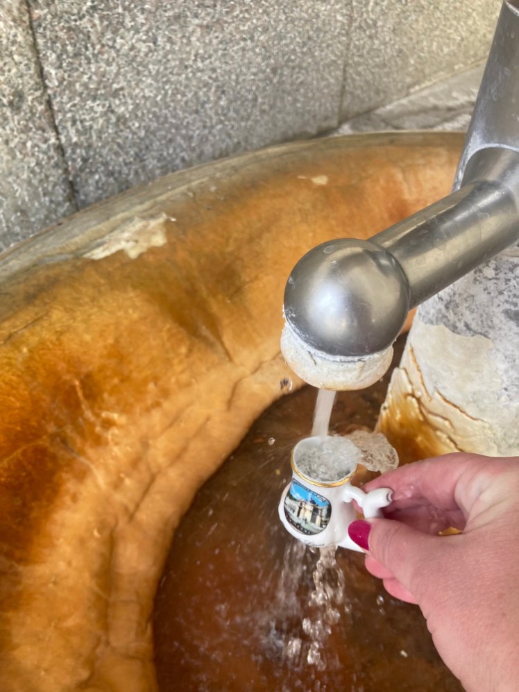 My tiny spa cup overflowing as I hold it under a spout. The basin underneath is thick with orange mineral build-up.