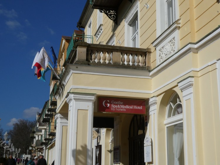 A yellow and white hotel with balcony, flags, columns and all the decorative mouldings. Hanging from the porch is a sign declaring it "Goethe Spa & Medical Hotel".