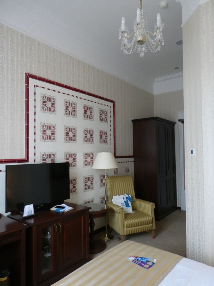 My room in the Nové Lázně. It's a small room but it's not tiny. It has original 1896 red and white tiles on the walls, a crystal chandelier, a proper yellowish armchair and heavy furniture in dark brown wood.