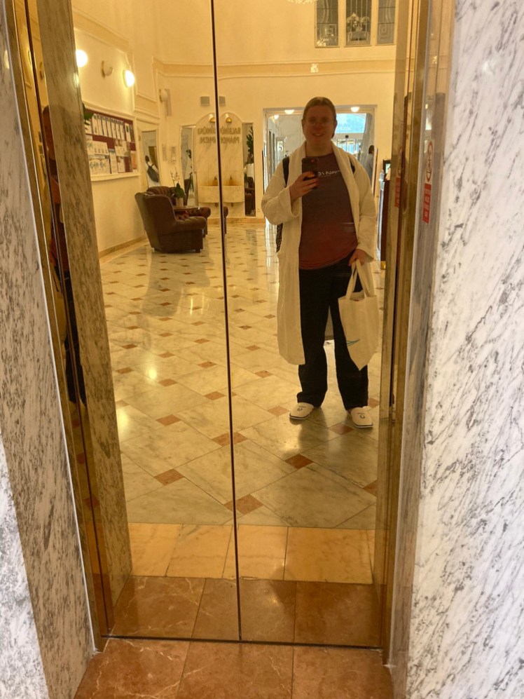 A selfie in black hiking trousers, a purple t-shirt, spa robe and spa slippers in the gold reflection of the lift in the Hvězda hotel lobby.
