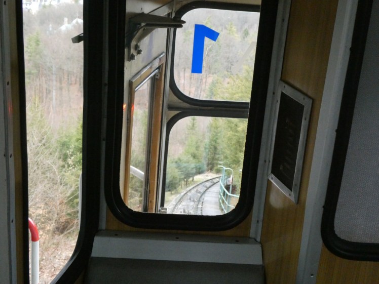 Inside the funicular, looking down at the narrow window between the section below me and the driving window, to see the track winding its way up the tree-covered hillside.