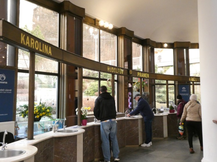 A row of sinks in the pavilion. There's a wooden display over them with the names in big gold letters - Karolina, Rudolfův and Křížový.