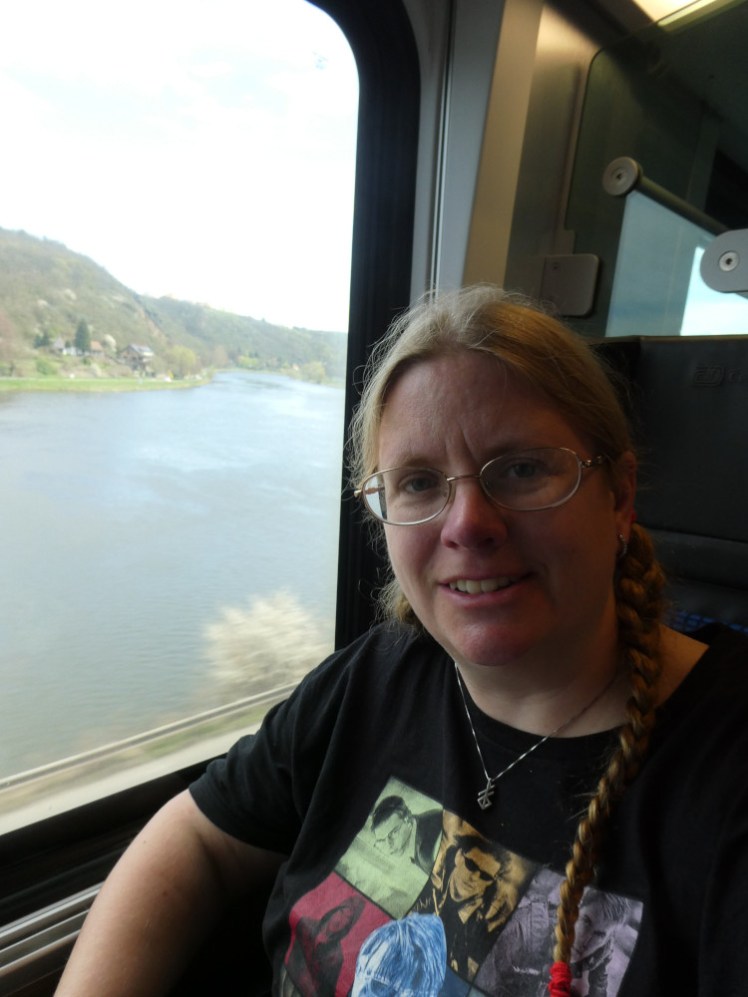 A selfie on the train to Prague, wearing my Ethan Hunt t-shirt. Behind me, you can see a wide river with green hills rising up out of it.