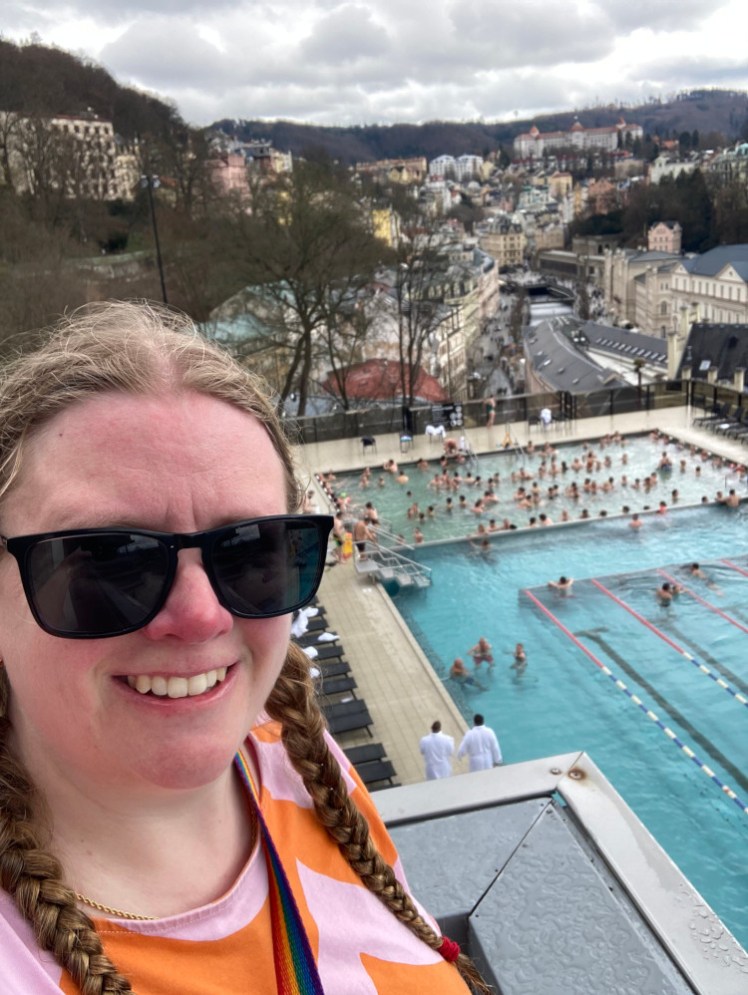 A selfie taken from the ledge above the pool. I'm wearing a pink and orange scalloped striped t-shirt and my hair is wet because I've just been in the pools.