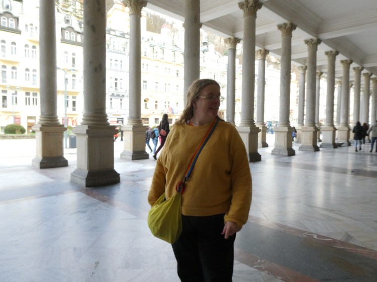 A selfie in the Mill Colonnade, wearing a yellow jumper and with my hair down for once. Behind me are a double row of stone columns and behind that, you can see the street of Karlovy Vary basking in the sun.
