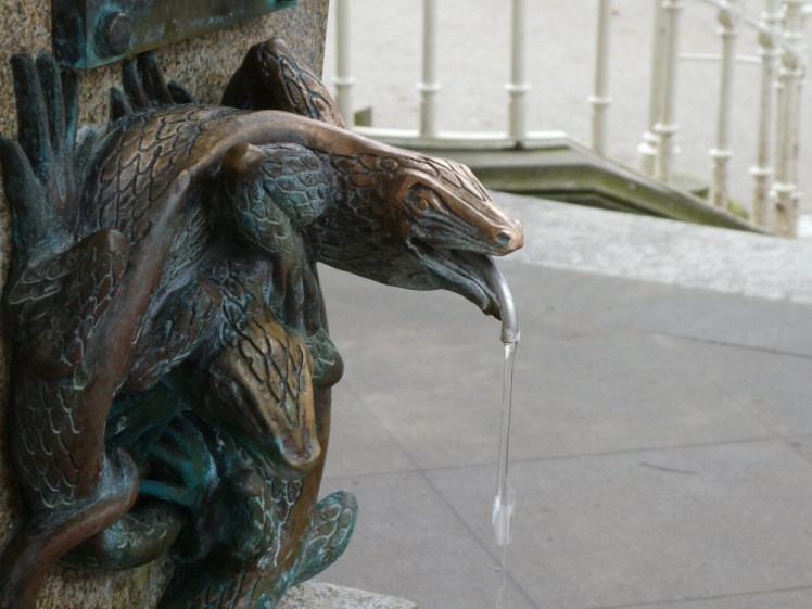A close-up of a tap spouting hot spring water. The tap is bronze and shaped like a snake peering out of a tangle of snakes.