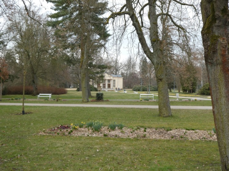 The gardens in Františkovy Lázně, dotted with trees and flower beds and criss-crossed with paths and benches. Through the trees, you can see a classical-style building which houses another spring and several sinks for tasting it.