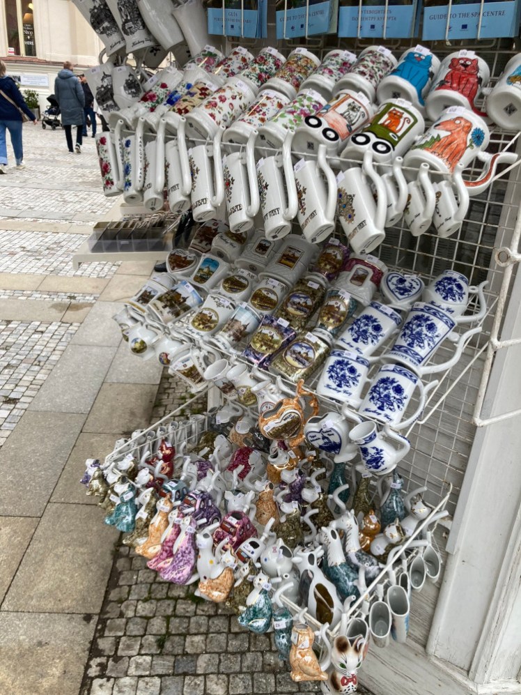 An assortment of spa mugs hanging outside a kiosk. The ones at the top are illustrated, there are flat ones hanging from the top rack, small ones hanging from the middle rack and the entire bottom rack is animal-shaped cups.