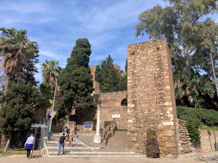The main entrance to the Alcazaba, a stone tower and stone walls going backwards and forwards up the hill, half-hidden behind trees.