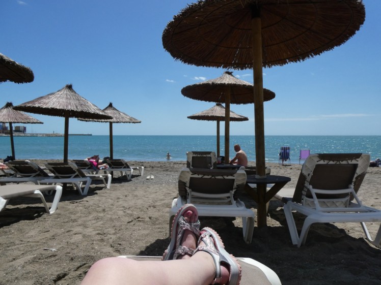 My feet in sandals lying on a sunlounger on a grey-brown beach under thatched parachutes with the sun and sea looking so perfect you'd never guess there's a strong and freezing wind.