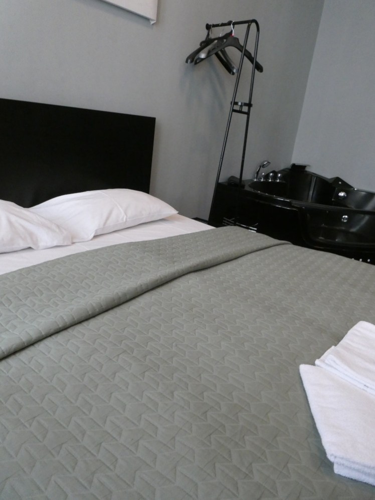 The bed, with a grey cover on it, a wire rack next to it and the bath on the other side of the rack.