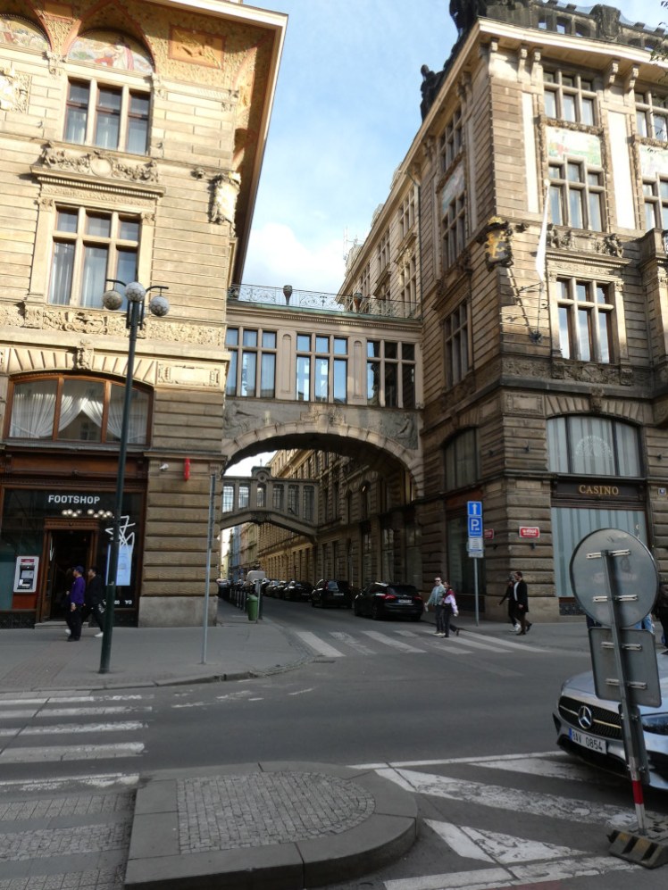 A street corner with two buildings on the other side of the road, joined by a bridge with windows in it. There are zebra crossings all around it. This is where Ethan was picked up in the big car with the shroud.