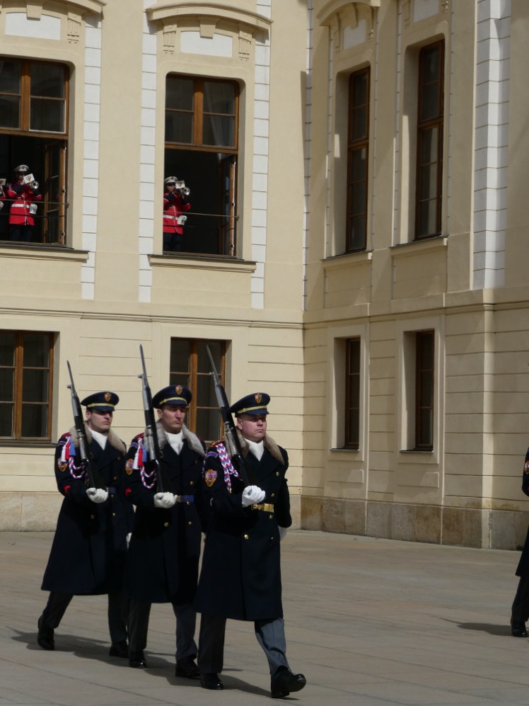 Some of the guard marching across the courtyard with rifles and fur collars  and military musicians in the windows above dressed in red uniforms.