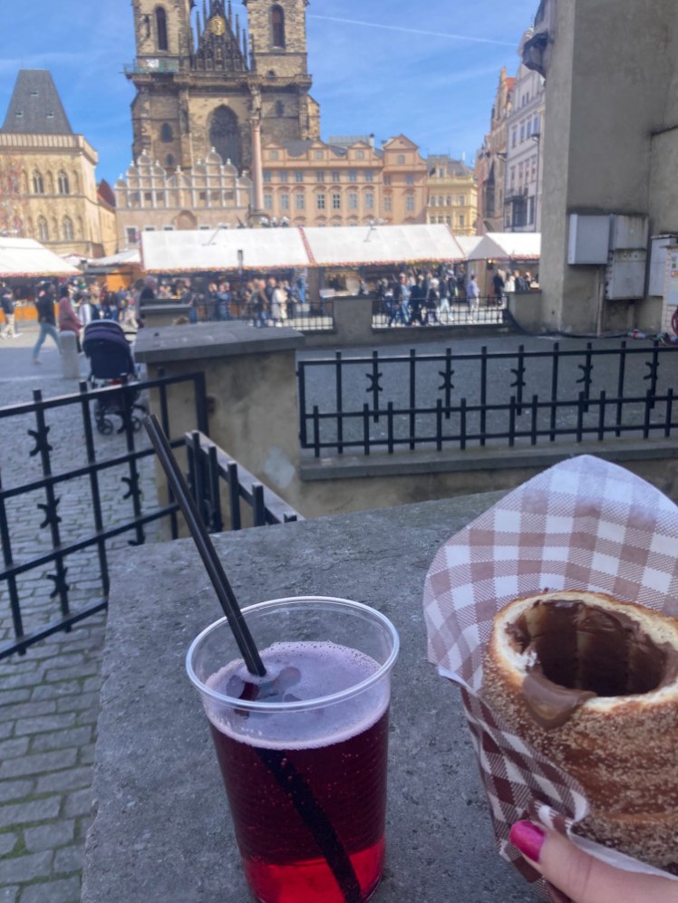 A plastic cup of dark pink raspberry lemonade and a chimney cake in checked paper with the Old Town Square and Easter market in the background.