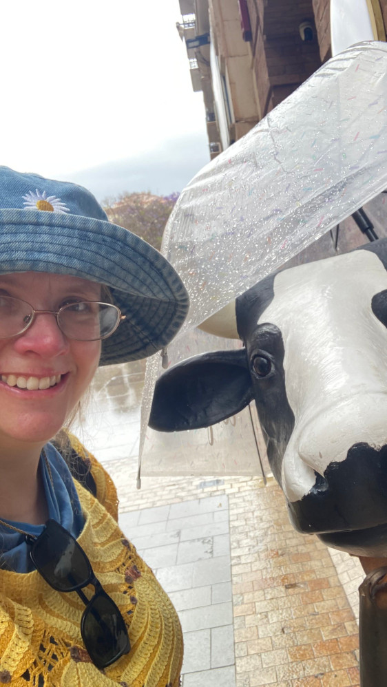 A selfie in the rain with a fibreglass cow with an umbrella on its head.