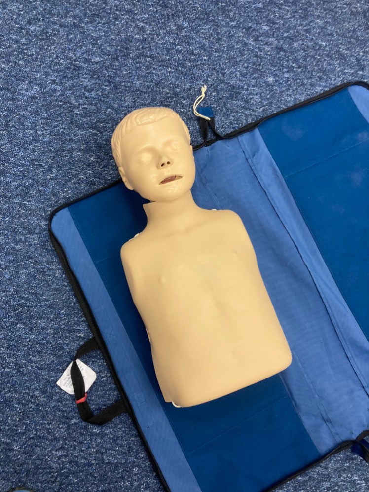 A CPR mannequin lying on its unfolded bag. This one is a junior model, a child dummy, a torso and head with a dead lipless face and no limbs.