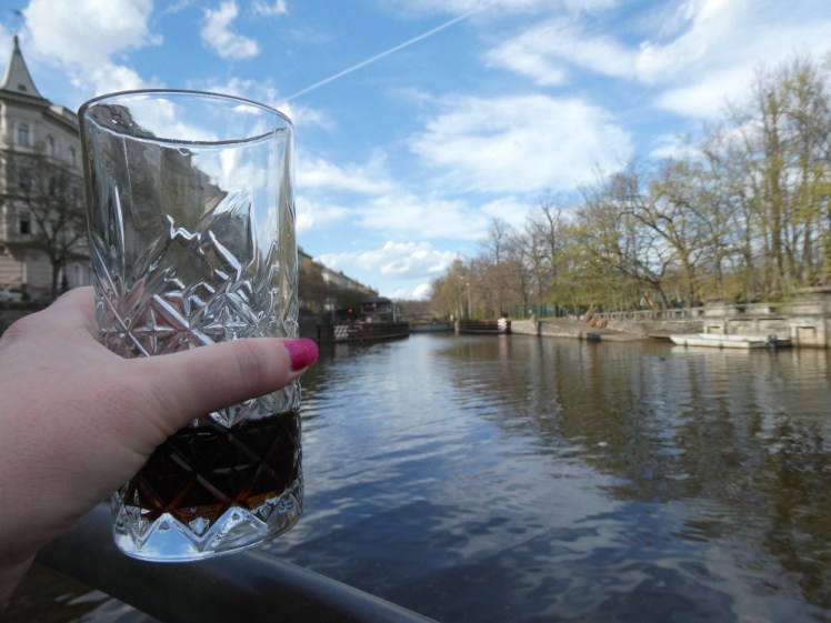 Me hand holding out a decorative tumblr with two fingers of Coca-Cola in the bottom, held above the river under a blue sky.