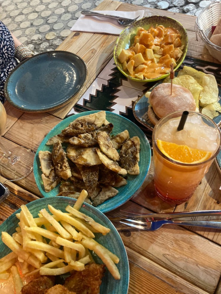 A table of tapas, including potatoes with spicy sauce, a mini burger, fried aubergines in honey and some breaded meat thing with chips, as well as some bright yellow fruity juice drinks.