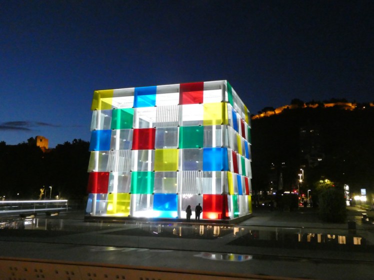 The Centre Pompidou by night, a house-sized cube of coloured glass with the Gibralfaro illuminated on the hill behind it.