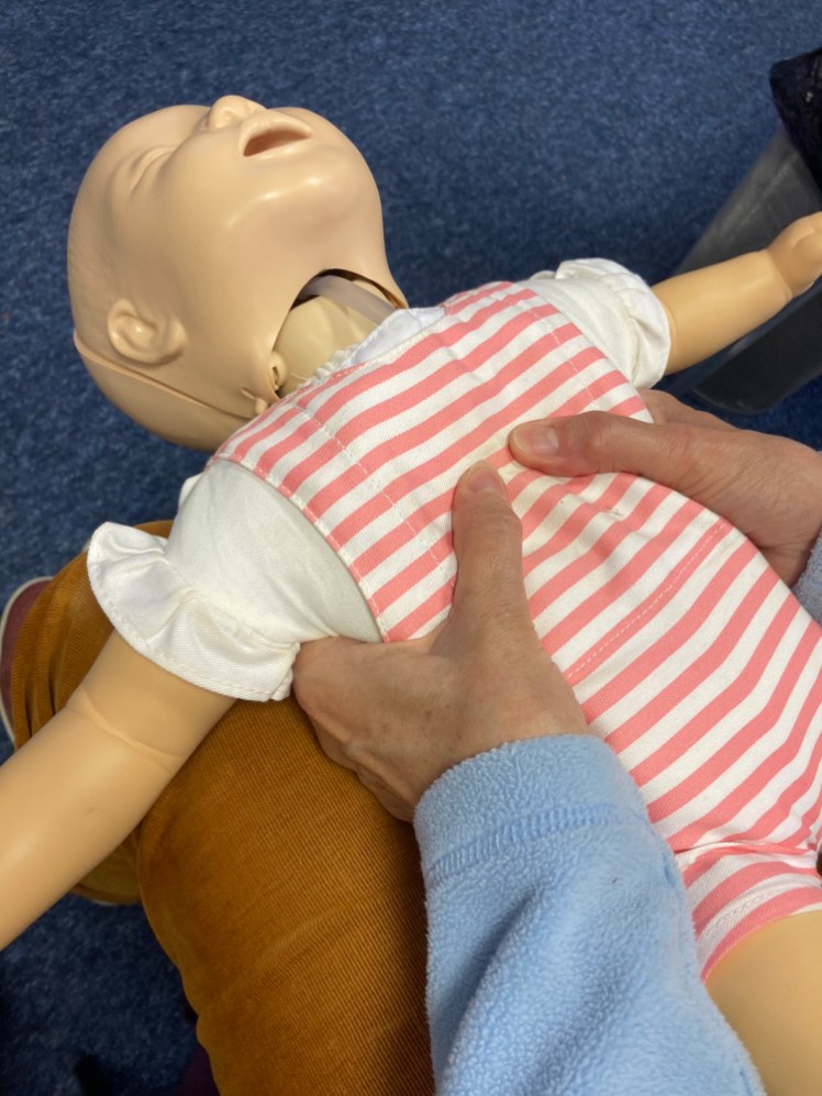A baby manneqiun held on someone's knees, on its back, with their hands around its chest and thumbs pressed in.