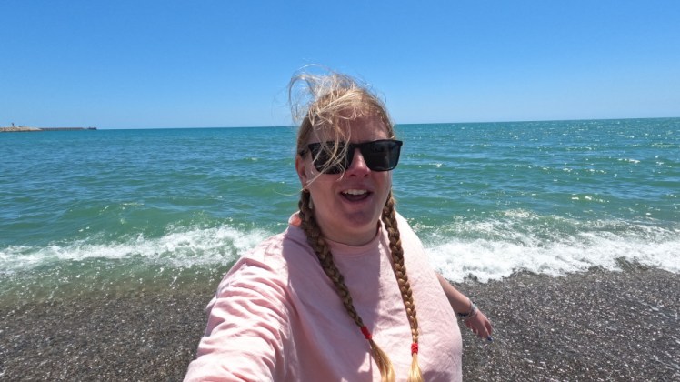Me on the beach, looking a bit windswept in a pale pink t-shirt with a vivid blue sky and an incredible coloured Mediterranean sea behind me.