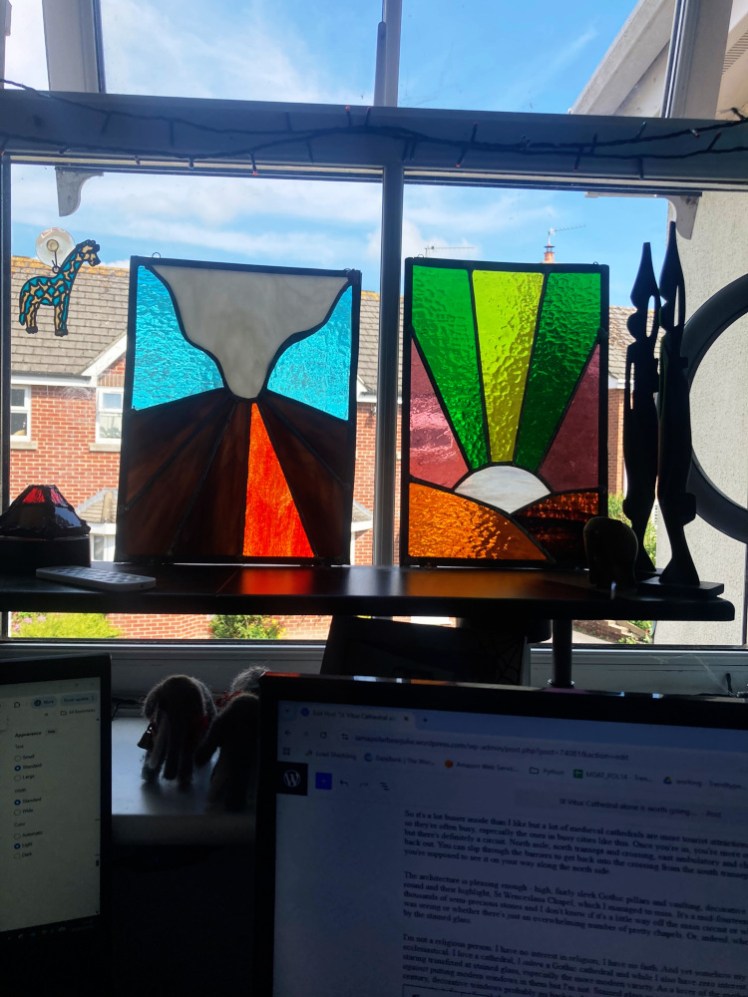 Two stained glass panels depicting a volcano and northern lights, in a vaguely Art Deco style, propped up on a little shelf on my windowsill. Below them, you can see this blog post in progress.
