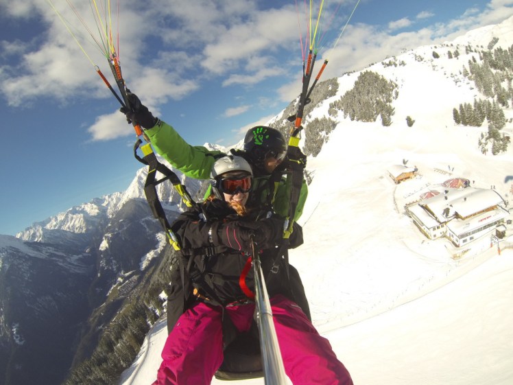 A picture of me in my hot pink snowboarding trousers and black jacket tandem paragliding over a snowy mountain in Austria. This is on a GoPro with a really thick pole right down the middle of the picture.