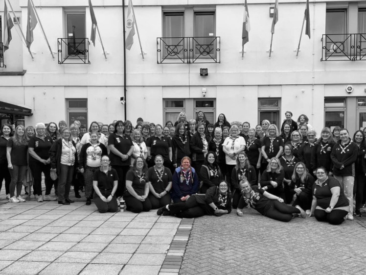 A group photo of leaders from Girlguiding South West gathered in uniform in front of Pax Lodge. It's all in black and white except me, in colour, towards the front.