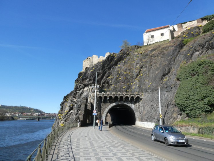 The Ruins of Libuses's Bath on a cliff with a road drilled through it, the river to the left and the blue sky over it all.