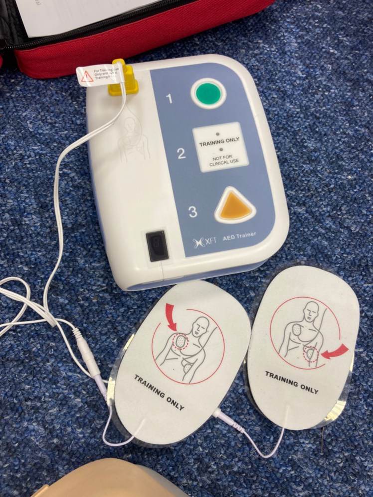 A training defibrillator on the floor, attached to a pair of sticky pads. There's a green button to activate it and a yellow triangular button to  administer a shock (except it's for training and it doesn't do anything except talk).