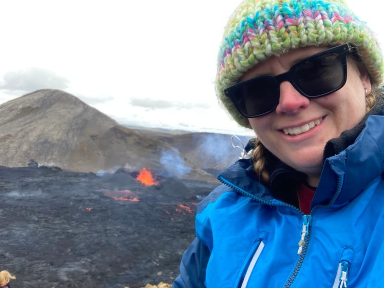 A selfie in blue raincoat and pastel rainbow striped knitted hat despite it being August, with a small but actively erupting volcano in the background.