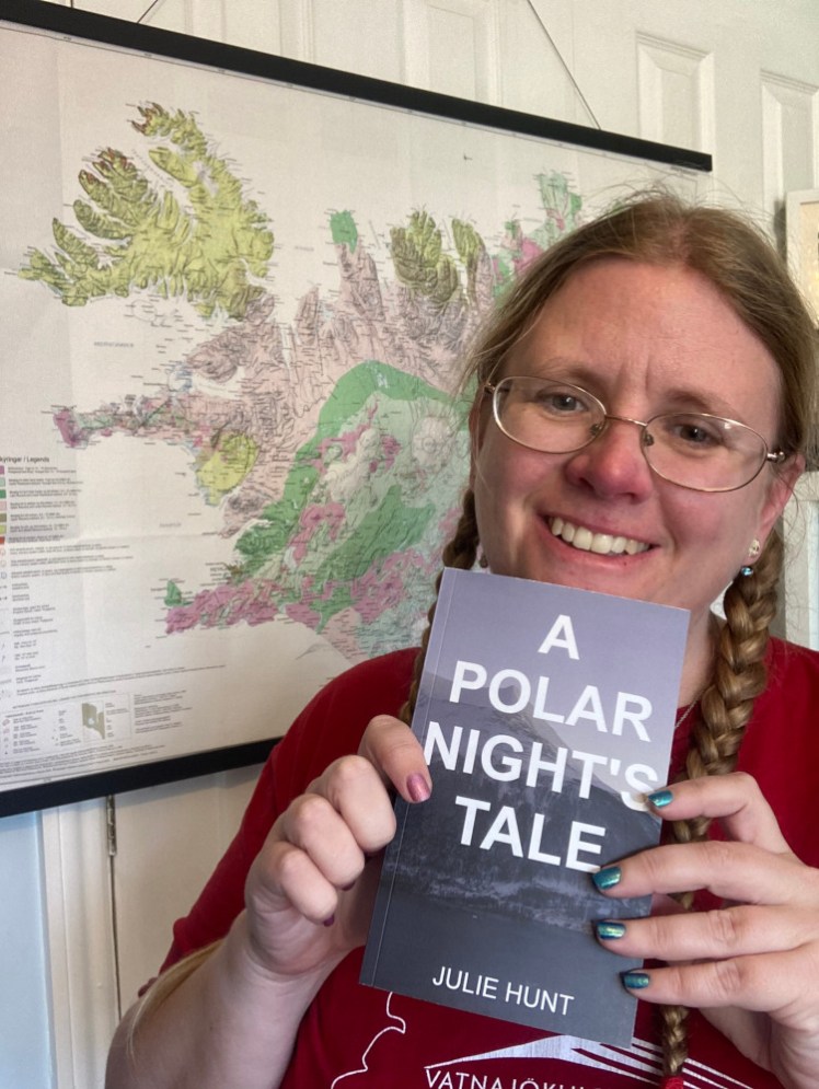 Me, next to my Iceland geological map, holding up my book, A Polar Night's Tale which has its title in big white letters over a picture of a mountain on a blue day in Norway.