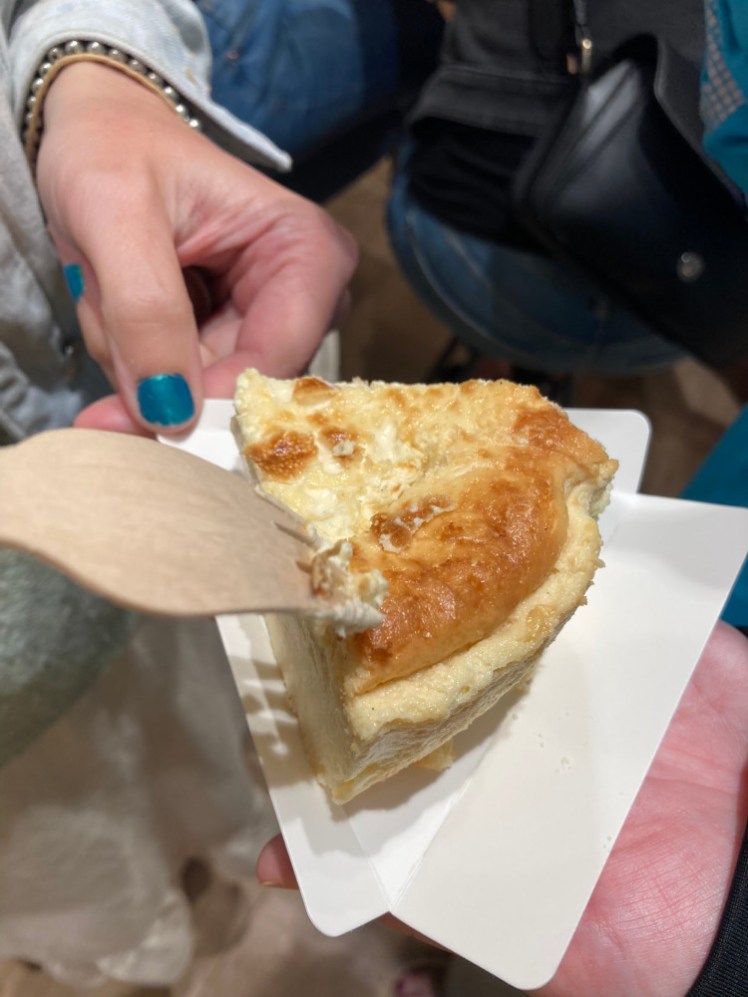 A wedge of mousse-like caramelised Basque-style cheesecake on a triangular paper plate. Catherine is just sticking a fork in it as I take a photo of it.