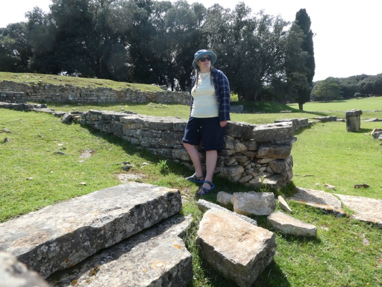 Me leaning on a Roman ruin in Croatia, pretending to look off into the distance. I'm wearing shorts (actually linen-mix trousers rolled up), a pale yellow t-shirt under a blue & white checked shirt, a bucket hat and my blue children's sandals.