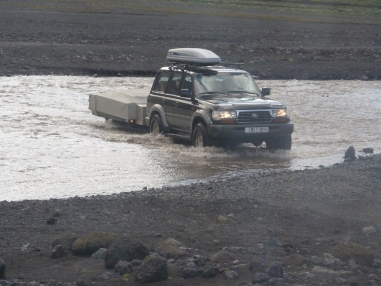 A 4x4 with a trailer in water that's coming up over the upper half of its wheels.